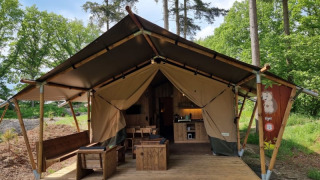 Tienda safari con servicios sanitarios, terraza de madera y muebles, situada en camping junto al bosque.