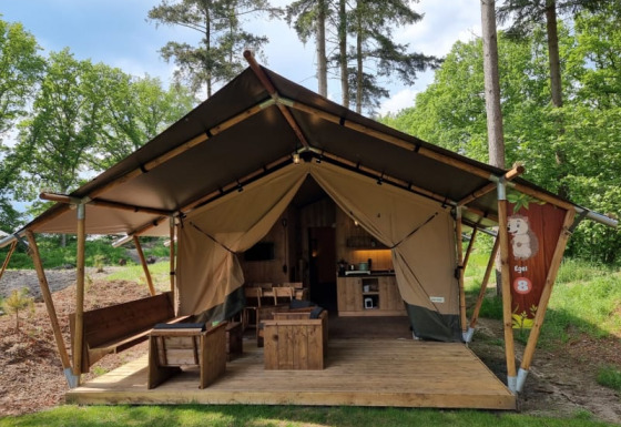 Tienda safari con servicios sanitarios, terraza de madera y muebles, situada en camping junto al bosque.