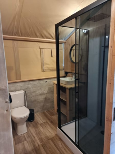 Bagno in tenda safari con box doccia in vetro, wc, lavabo in legno e illuminazione naturale.