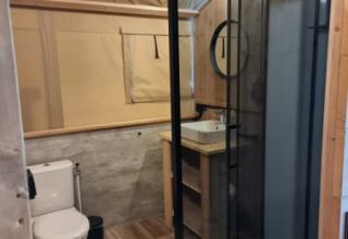Salle de bain sous tente safari avec douche vitrée, toilettes, vasque bois et éclairage naturel.