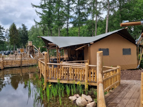Tienda safari con instalaciones sanitarias en terraza de madera junto a un estanque y bosque verde.