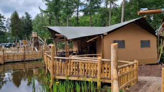Tienda safari con instalaciones sanitarias en terraza de madera junto a un estanque y bosque verde.