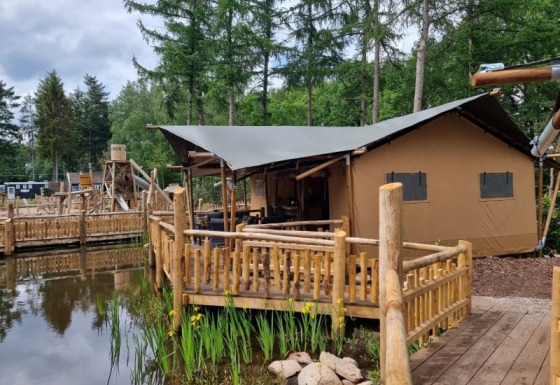 Tienda safari con instalaciones sanitarias en terraza de madera junto a un estanque y bosque verde.