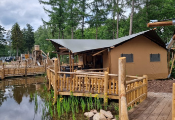 Safari tent with private sanitary facilities on wooden deck by a pond, surrounded by lush green forest.