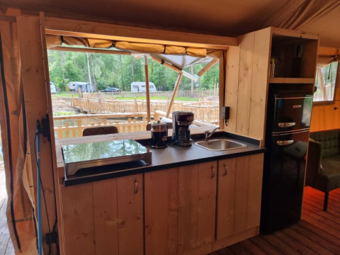 Área de cocina en tienda safari con muebles de madera, cafetera, fregadero y vistas al campamento.