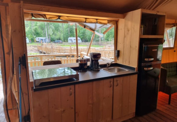 Área de cocina en tienda safari con muebles de madera, cafetera, fregadero y vistas al campamento.