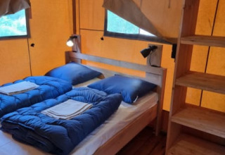 Vista interna di una tenda safari con due letti singoli, biancheria blu e pavimento in legno al Camping het Horstmannsbos.