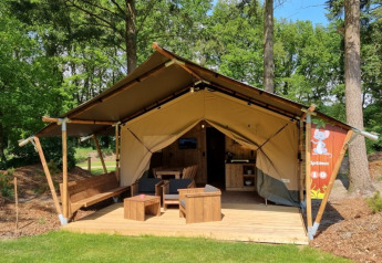 Tenda safari con servizi igienici privati, veranda in legno e zona relax all'aperto in ambiente boschivo.