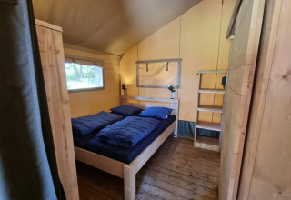 Interior view of a safari tent with wooden floor, double bed, blue bedding and rustic wood furniture.