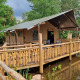 Safari-teltet Safari tent + sanitary - Baars med en stor træterrasse ved en sø, omgivet af grøn skov.
