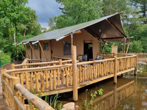 Safari tent called Safari tent + sanitary - Baars, with a spacious wooden deck over water in a green forest.