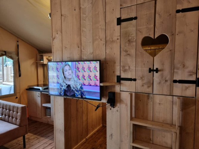 Interior de tienda safari con paredes de madera, televisor y recorte de corazón, Camping het Horstmannsbos.