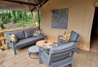 Zona salotto esterna con mobili grigi e cuscini floreali davanti a una tenda safari al Camping het Horstmannsbos.