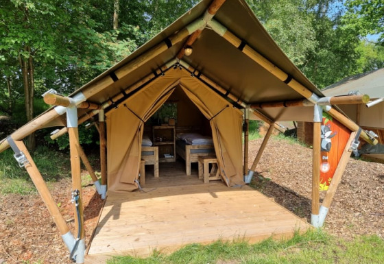 Safari tent named Duolodge with wooden deck and beds, set on a campsite surrounded by green trees.