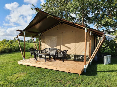 Safari tent with wooden deck and plastic chairs, set in a grassy area with trees under a blue sky.