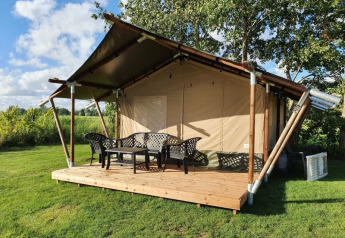 Tienda safari con terraza de madera y sillas plásticas, rodeada de césped y árboles bajo cielo azul.