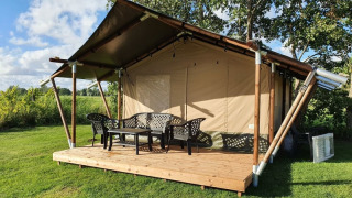 Tente safari avec terrasse en bois et chaises en plastique, entourée de pelouse et d’arbres sous ciel bleu.