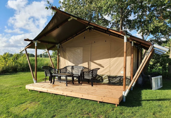 Safari tent with wooden deck and plastic chairs, set in a grassy area with trees under a blue sky.