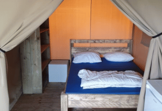 Vista interior de una tienda safari con suelo de madera, cama doble, estanterías y un pequeño frigorífico.