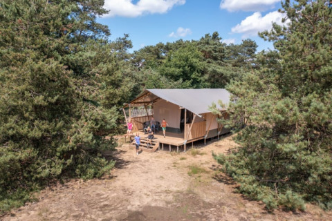 Safari-Zelt mitten im Wald mit Menschen auf der Veranda, umgeben von Bäumen und blauem Himmel.