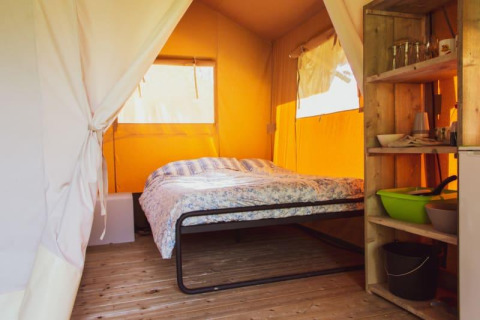 Interior de una tienda safari con cama, estantería de madera y básicos de camping en suelo de madera.