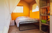 Interior de una tienda safari con cama, estantería de madera y básicos de camping en suelo de madera.