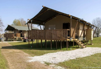 Safari-Zelt mit sanitären Anlagen, steht erhöht auf Stelzen, Holzterrasse, grüner Rasen und blauer Himmel.