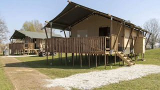 Tente safari avec sanitaires, sur pilotis, terrasse en bois, pelouse verte sous ciel bleu dégagé.