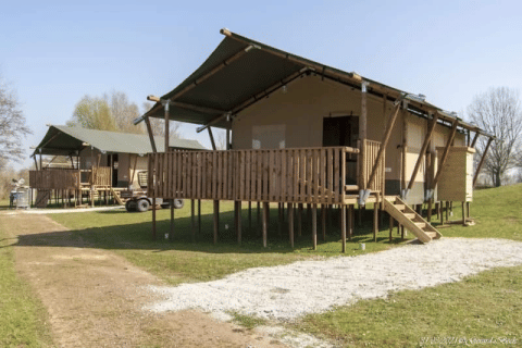 Safari tent with sanitary facilities, raised on stilts, wooden deck, set on grassy land under a blue sky.