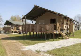 Tente safari avec sanitaires, sur pilotis, terrasse en bois, pelouse verte sous ciel bleu dégagé.