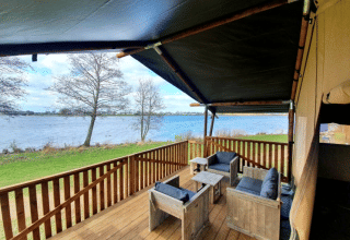Terraza cubierta con muebles de madera y vistas al río en Safari tent + sanitary, Recreatiepark Riverside, Países Bajos.