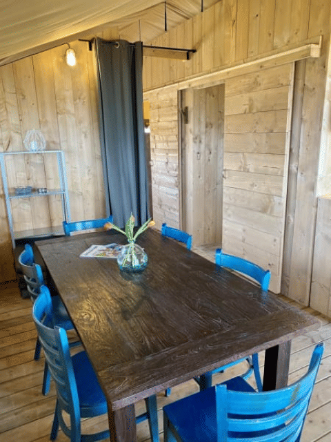 Zona pranzo in una tenda safari con pareti di legno, tavolo scuro, sedie blu e vaso di fiori in Olanda.