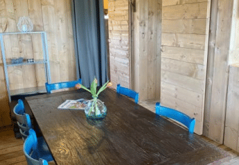 Comedor dentro de una tienda safari con paredes de madera, mesa oscura, sillas azules y jarrón con flores en Holanda.