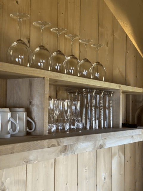 Wooden shelf in a safari tent kitchen with wine glasses and mugs at Recreatiepark Riverside, Netherlands.