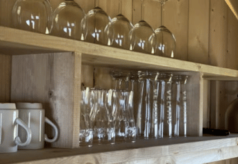 Wooden shelf in a safari tent kitchen with wine glasses and mugs at Recreatiepark Riverside, Netherlands.