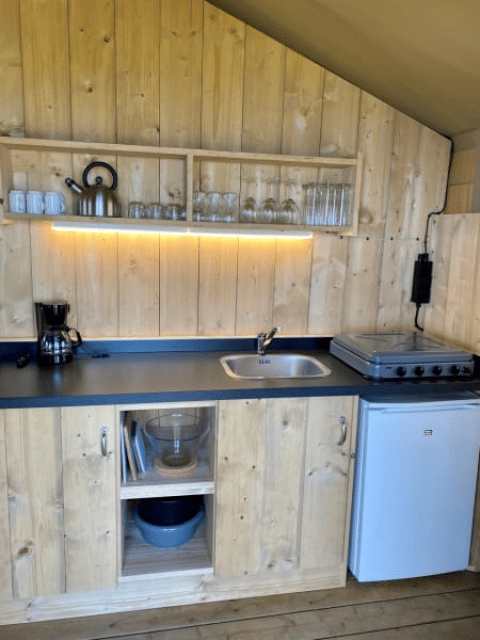 Kitchenette en bois dans une Safari tent + sanitary à Recreatiepark Riverside, Pays-Bas, avec cuisinière et réfrigérateur.