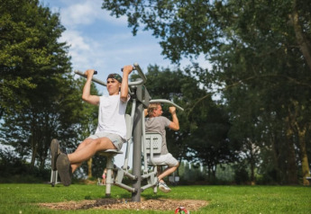 Due persone si allenano su attrezzi fitness all’aperto vicino a una tenda safari con servizi igienici al Recreatiepark Riverside, Paesi Bassi.