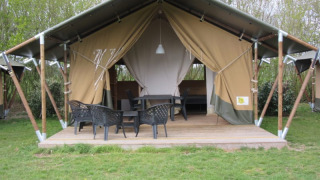 Safari tent with covered wooden deck, black chairs and table, surrounded by trees and green grass.