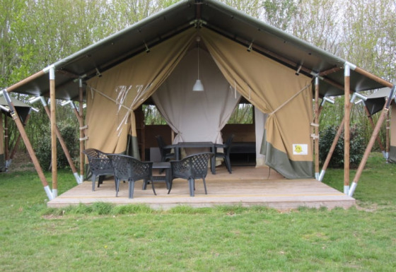 Tenda safari con veranda in legno, sedie e tavolo neri, circondata da alberi e prato verde.