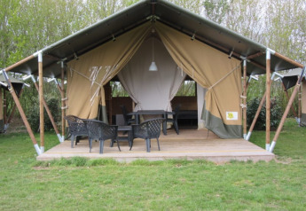 Safari tent with covered wooden deck, black chairs and table, surrounded by trees and green grass.