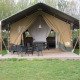Tienda safari con terraza de madera cubierta, sillas y mesa negras, rodeada de árboles y césped verde.
