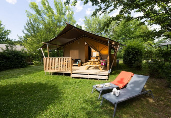 Safari tent met sanitair, houten veranda, picknicktafel en ligstoelen op zonnig grasveld omgeven door groen.