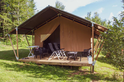 Safari-Zelt mit überdachter Veranda, Liegestühlen und Tisch, umgeben von Natur und Bäumen im Sonnenschein.