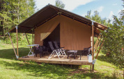 Safari tent with covered porch, lounge chairs, and a table, set in lush greenery and trees under blue sky.