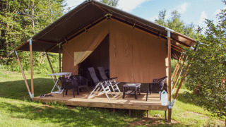 Tenda safari con veranda coperta, sedie sdraio e tavolo, immersa nel verde e circondata da alberi e cielo azzurro.
