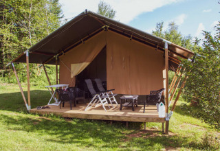 Safari tent with covered porch, lounge chairs, and a table, set in lush greenery and trees under blue sky.