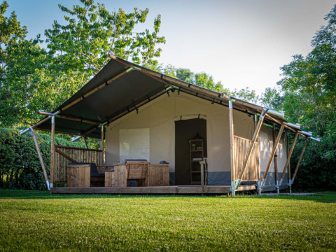 Safaritent met houten terras en buitenzitjes, gelegen te midden van grasveld en groene bomen.