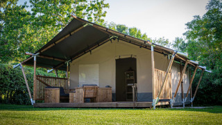 Safari tent with wooden porch and outdoor seating, surrounded by green grass and lush trees in nature.