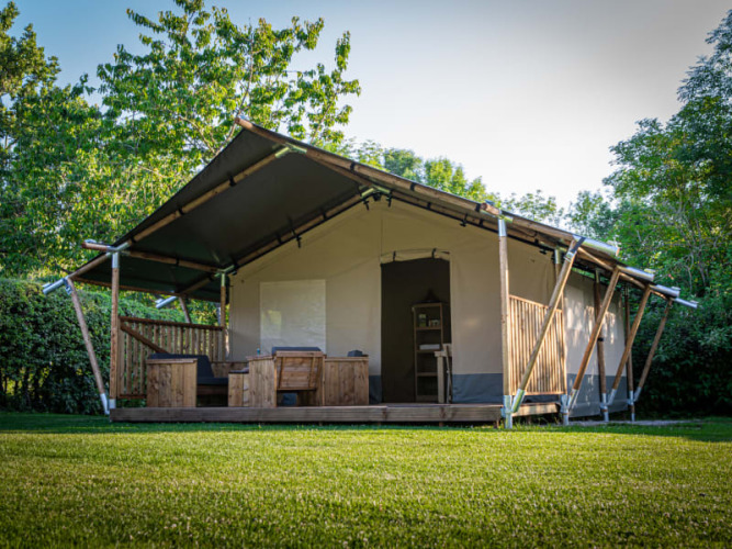 Safari tent with wooden porch and outdoor seating, surrounded by green grass and lush trees in nature.