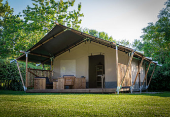 Safari tent with wooden porch and outdoor seating, surrounded by green grass and lush trees in nature.